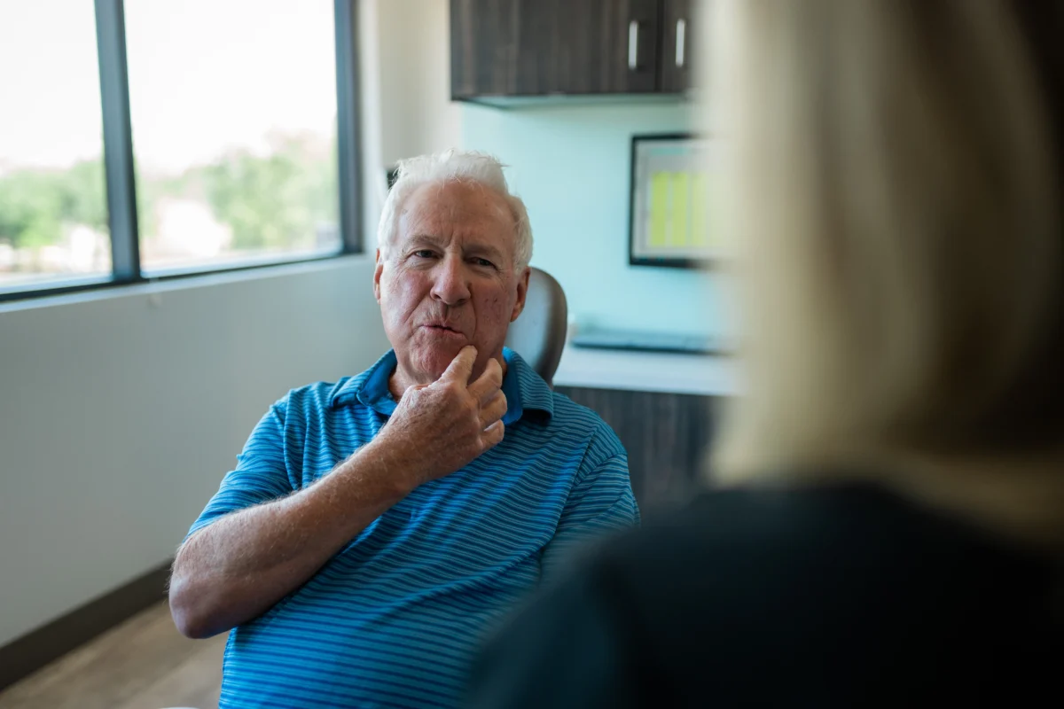 An older man in a blue striped shirt sits in a dental chair, touching his cheek while discussing a concern with a dental professional. The background features a modern dental office with natural lighting.
