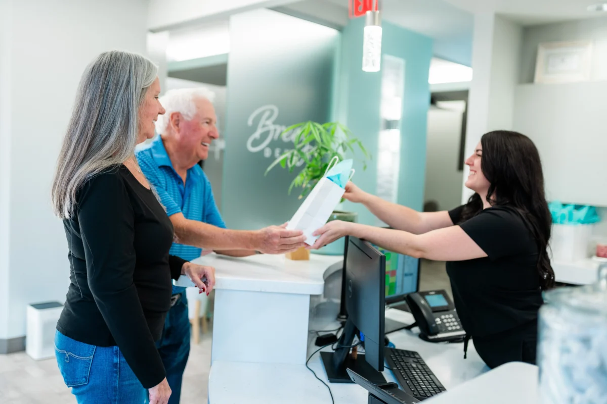 Receptionist at Breez Dental handing a gift bag to an elderly couple at the front desk